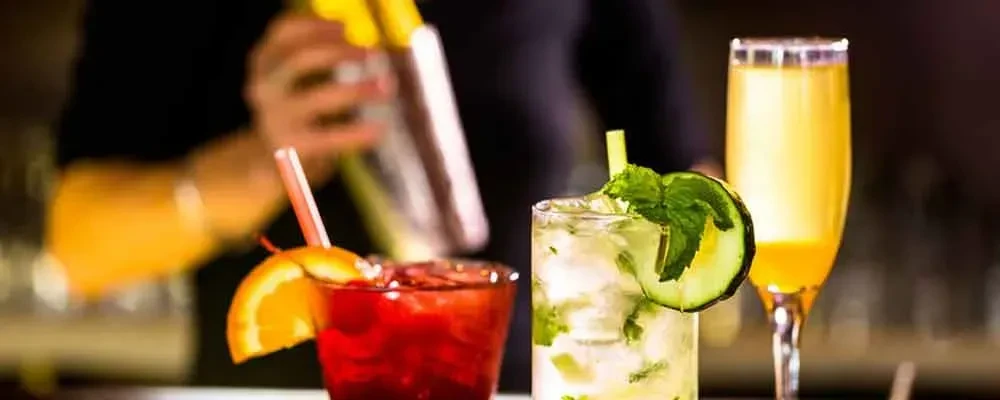 Three distinct cocktails on a bar, a bartender shakes a drink.