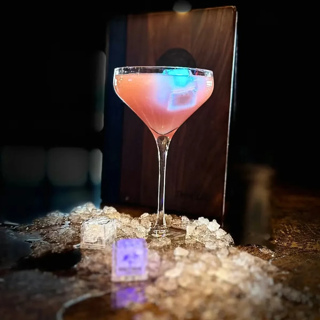 Pink cocktail in coupe glass, glowing ice cubes on crushed ice.
