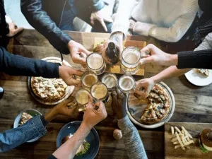 Overhead: hands toasting beer mugs with pizza and food on table.