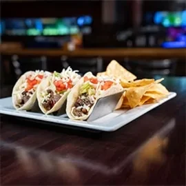 Three soft-shell tacos in a holder with meat, vegetables, and chips.