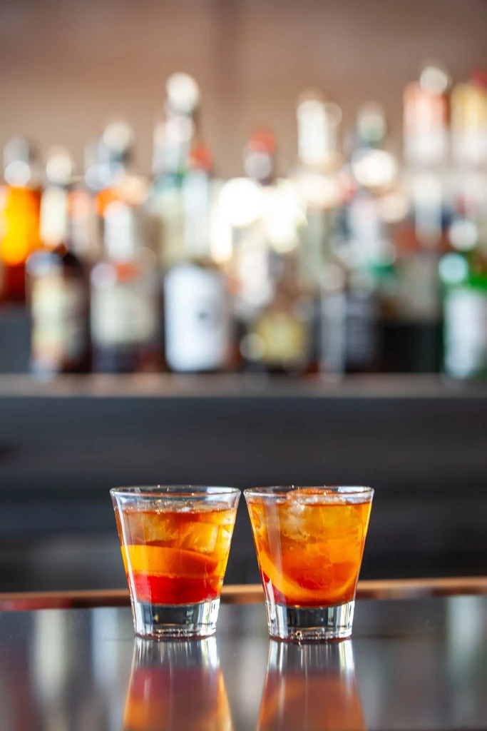 Twin amber cocktails with ice and citrus on a reflective bar.
