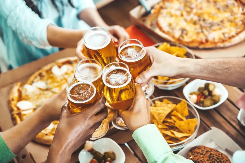 Five diverse hands clinking beer glasses above food.