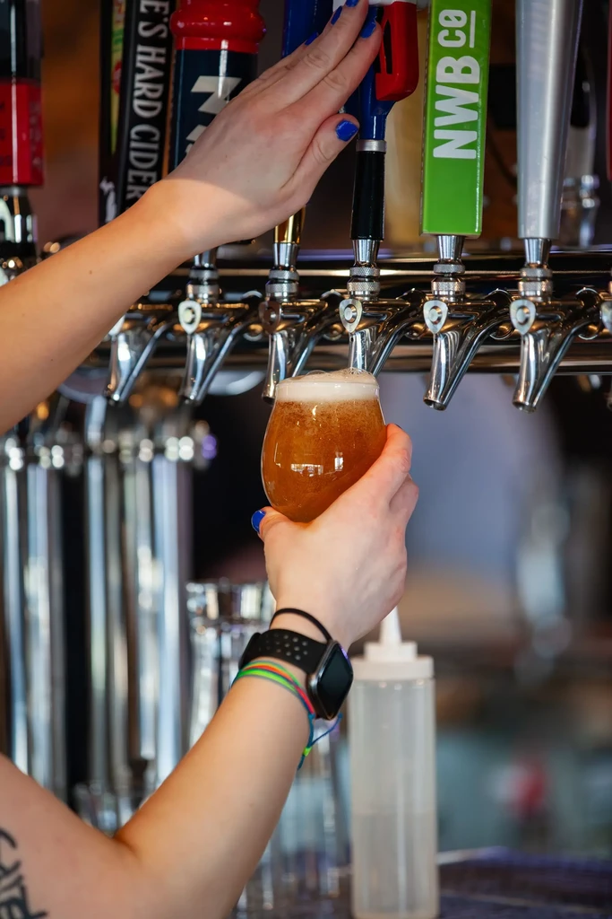Hands with blue nails pour frothy amber beer into a stemmed glass.