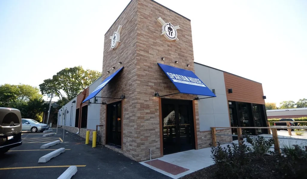 Modern Spartan House restaurant exterior, stone tower, blue awnings, parking lot.