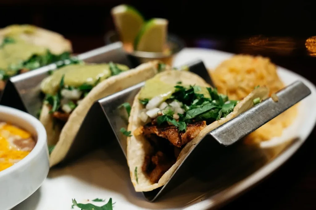 Three meat tacos in a stand, with beans, rice, and lime wedges.