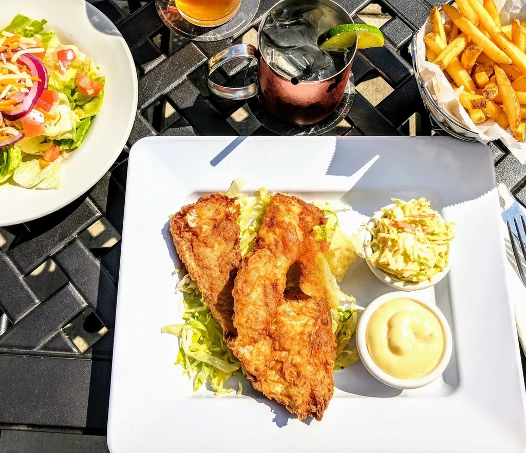 Crispy fried fish dinner with fries, salad, coleslaw, and drinks outdoors.