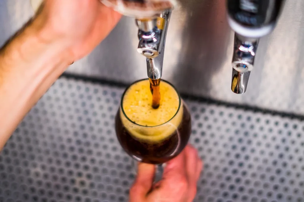 Hands pour frothy dark beer from chrome tap into stemmed glass.