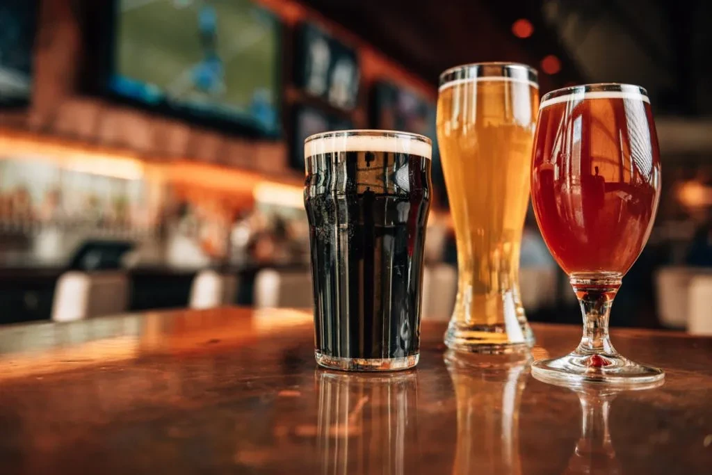 Three glasses of varied beers on a wooden bar with blurred TVs.