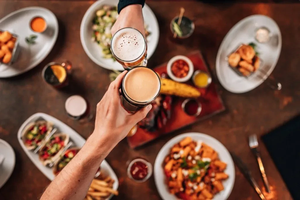 Two hands clink beer glasses surrounded by various dishes on a dark table.