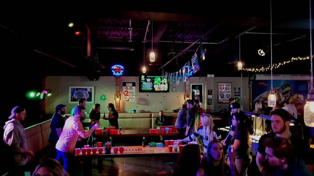 Dimly lit pub with people, neon signs, and a beer pong game.