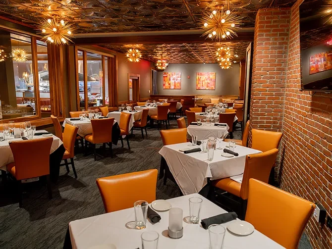 Upscale dining room with white tables, orange chairs, brick wall, and chandeliers.