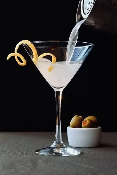 Cocktail being poured into martini glass with lemon peel and olives.