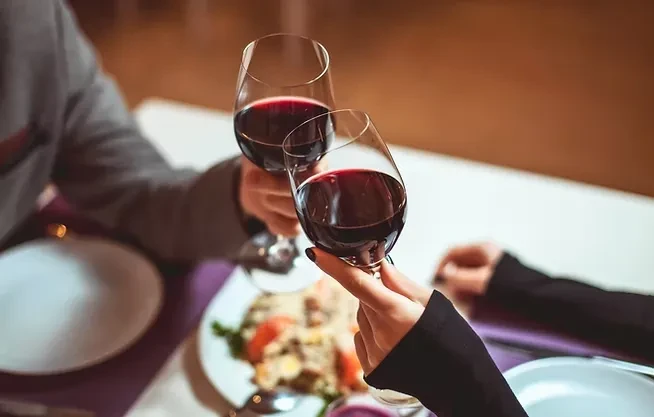 Two hands clink red wine glasses at a restaurant table.