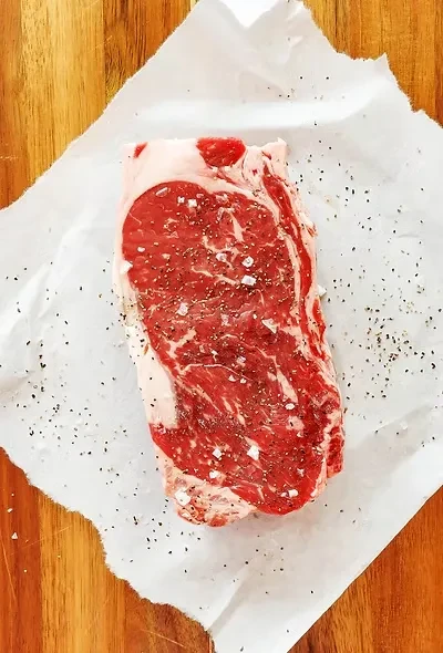 Raw, marbled steak with salt and pepper on paper and wood.