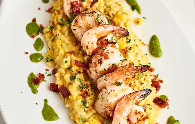 Close-up: Plated shrimp risotto, garnished with herbs, bacon, and green sauce.