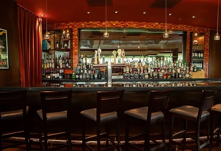 Empty bar area with six stools, glowing bottles, and an orange mosaic wall.