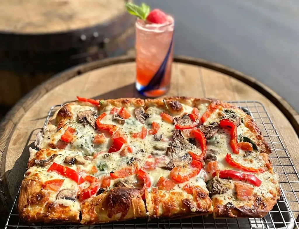 Rectangular pizza with toppings and a cocktail on a wooden barrel.