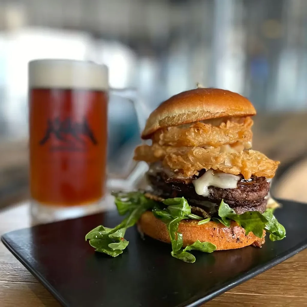 A close-up of a double-patty burger with onion rings, cheese, and beer.