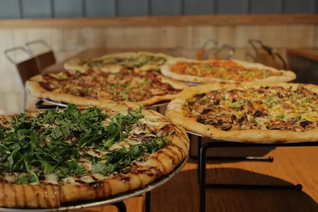 Five gourmet pizzas on tiered stands with varied fresh toppings.