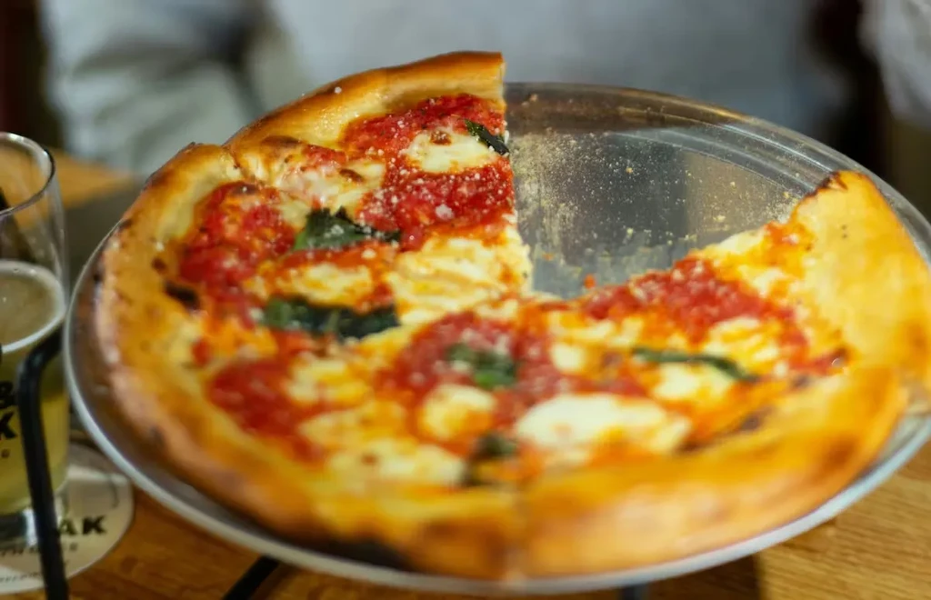Partially eaten Margherita pizza with basil and a beer on a wooden table.