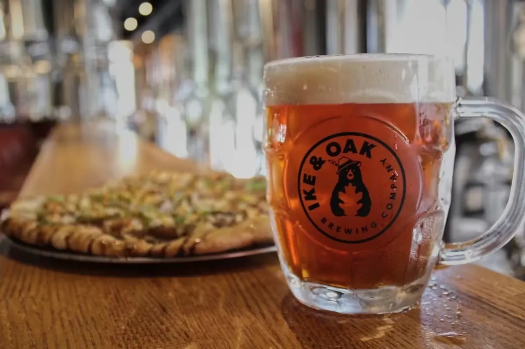 Pint of "IKE & OAK" amber beer with condensation and blurred pizza.