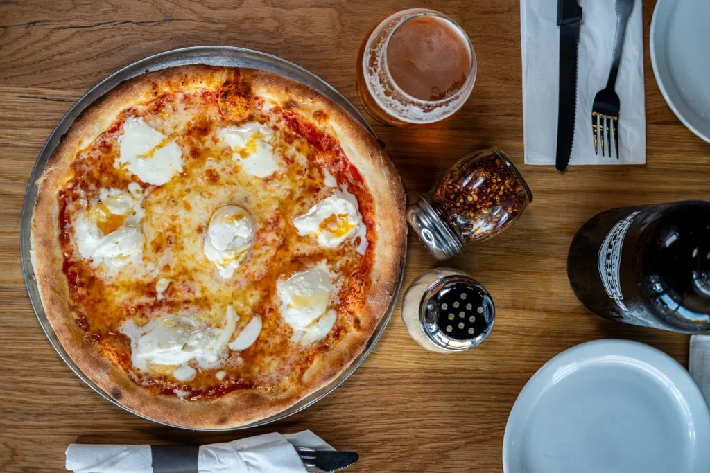 Pizza with melted cheese and burrata, beer, and condiments on a table.