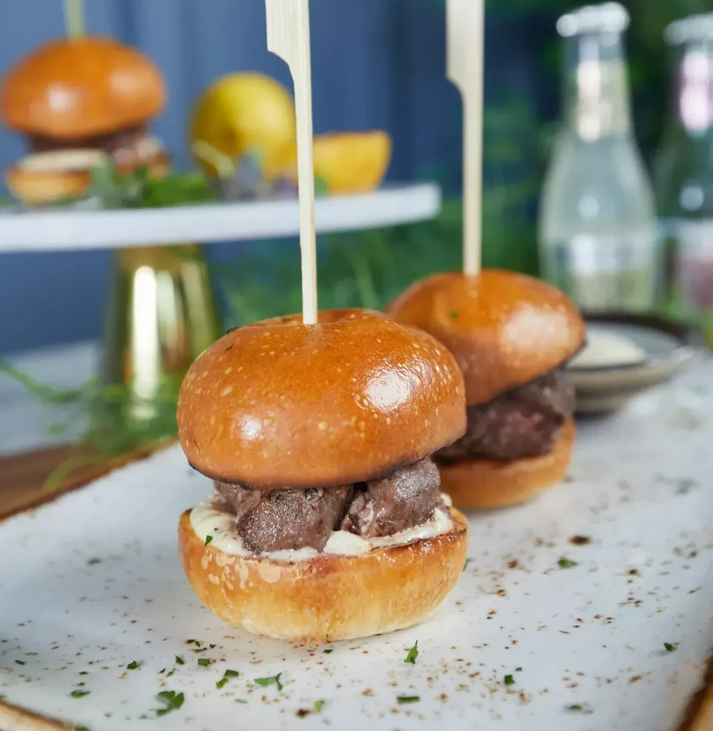 Two gourmet sliders with dark meat, sauce, skewers, and green herbs.