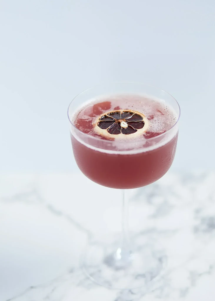 Reddish-pink cocktail in coupe glass, dried orange garnish on marble.