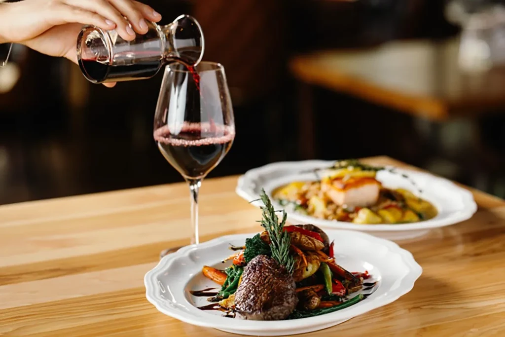 Hand pours red wine next to a well-presented meat and vegetable dish.