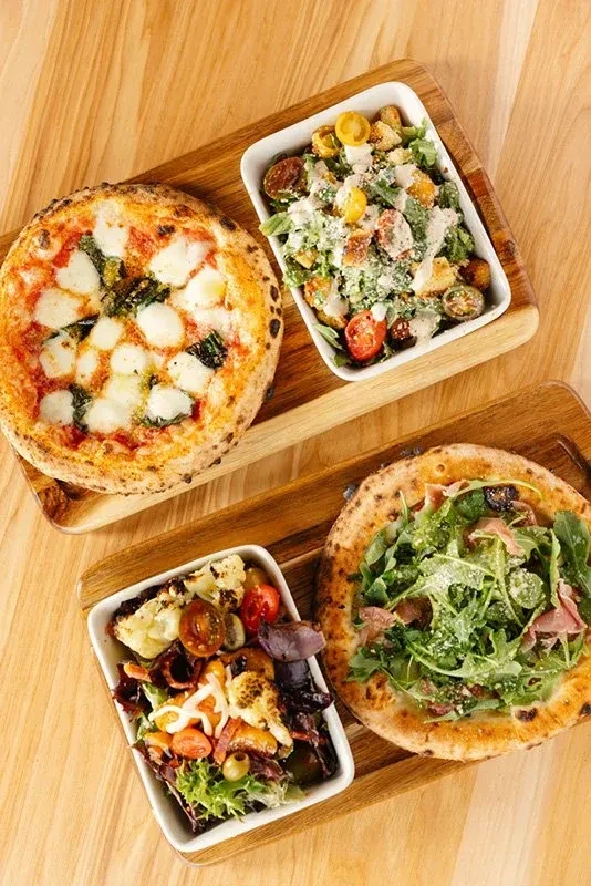 Overhead shot: two pizzas and two salads on wooden serving boards.