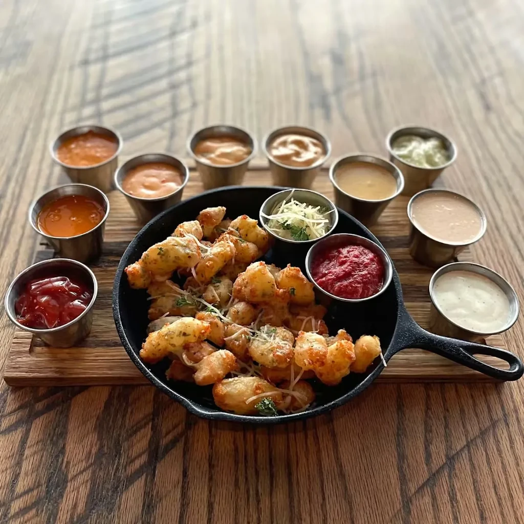 Cast-iron skillet of fried bites, ten assorted dipping sauces, wooden table.