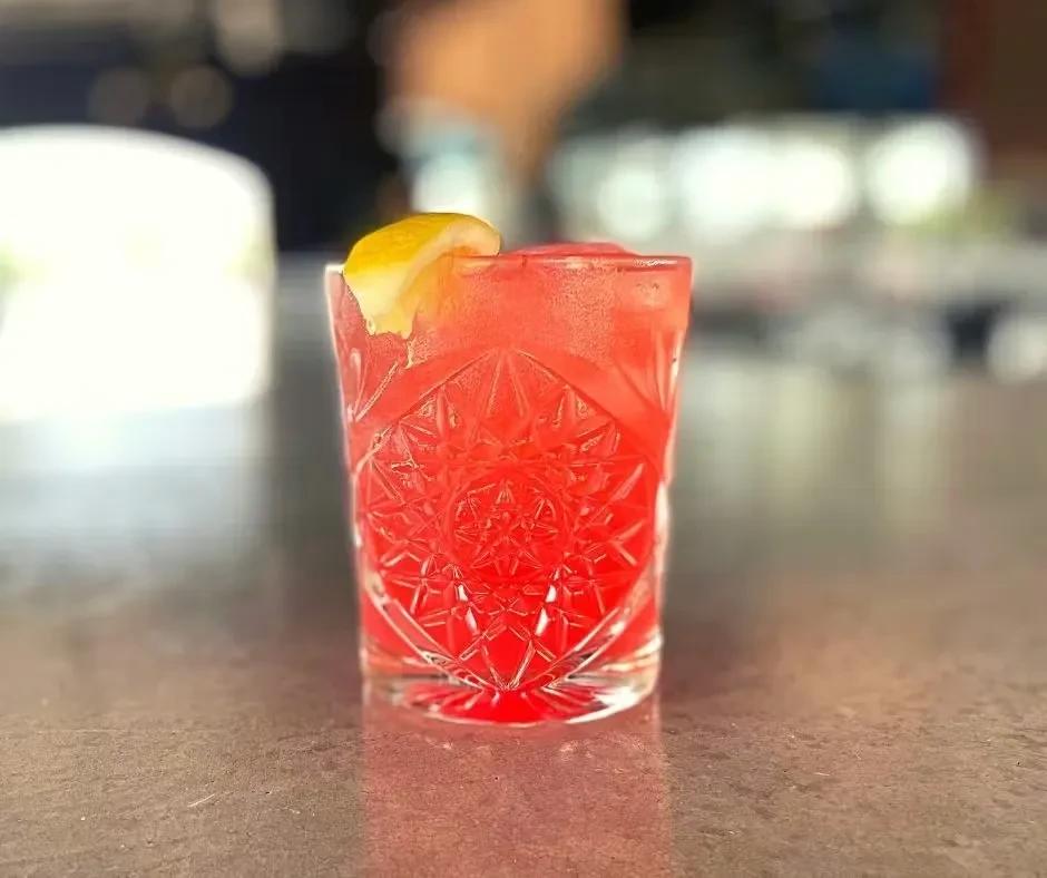 Cold red cocktail in cut-glass with lemon on dark counter.