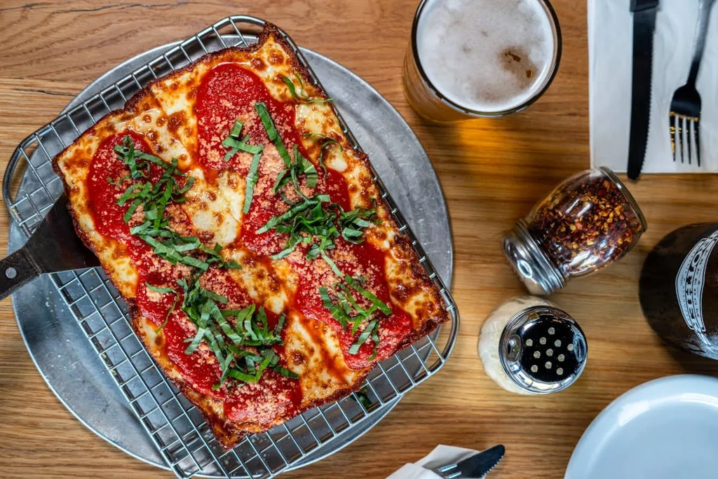 Rectangular Detroit-style pizza, beer, and condiments on a wooden table.