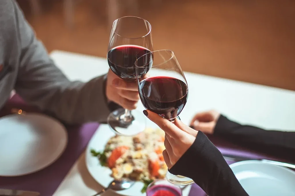 Two people toast with red wine glasses over a blurred dinner table.
