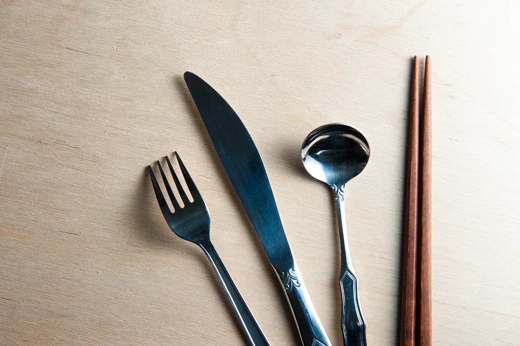 Silver fork, knife, spoon, and wooden chopsticks on light wood.