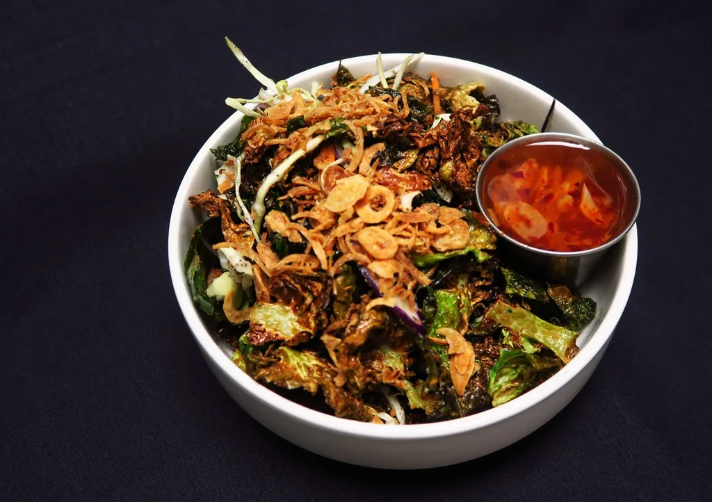 White bowl of roasted brussels sprouts, mixed greens, crispy onions, chili sauce.