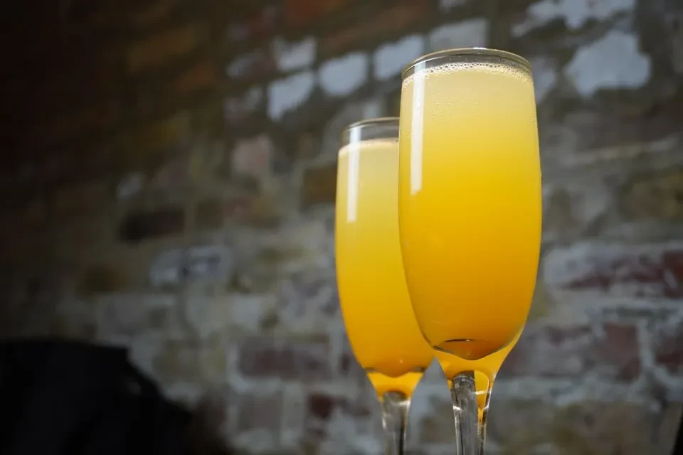 Two champagne flutes with orange beverages and foam against a brick wall.