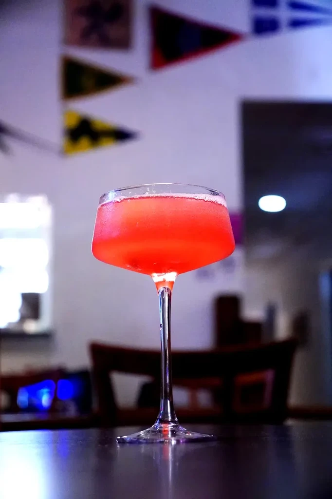Red or pink cocktail in a stemmed glass on a dark surface.