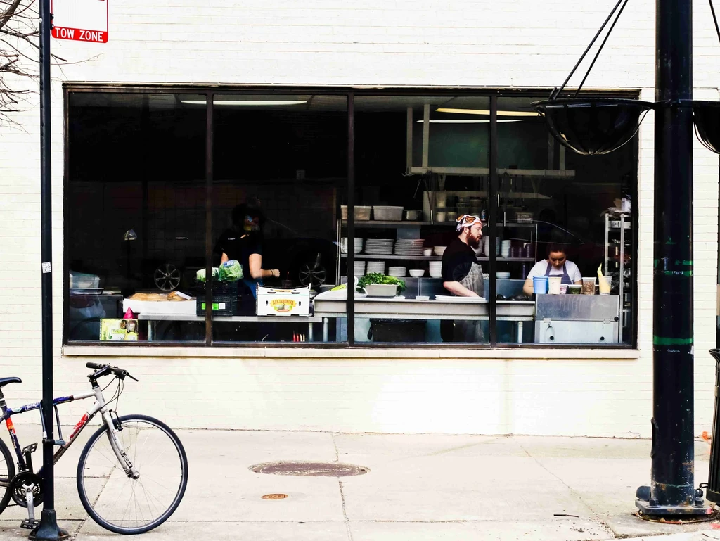 Restaurant kitchen viewed through window with staff, dinnerware, and equipment.