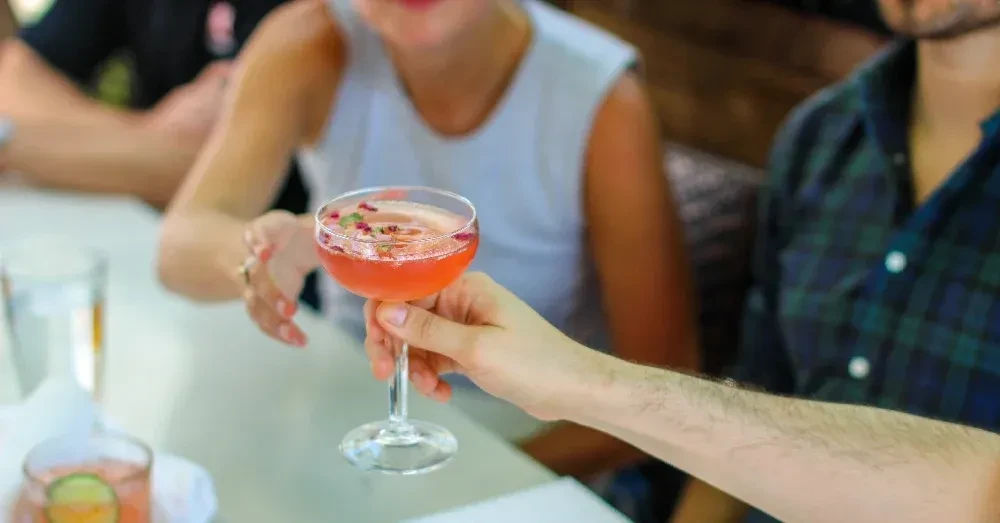 Hand holding a reddish-pink cocktail garnished with rose petals.