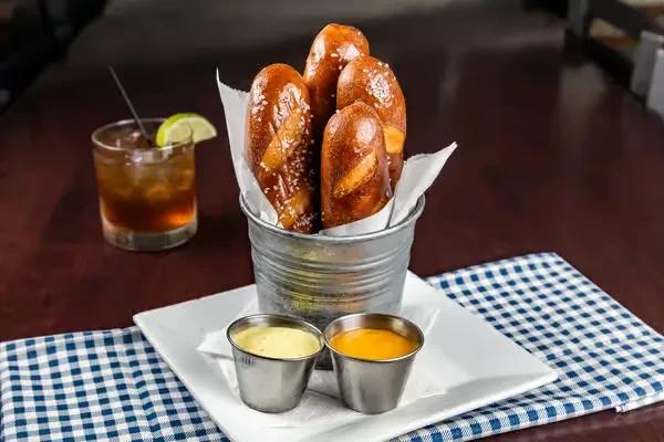 Pretzel sticks, honey mustard, cheese sauce, and a drink on a table.