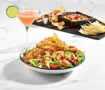 Chicken tender salad, fries, cocktail, chips, and queso on marble surface.