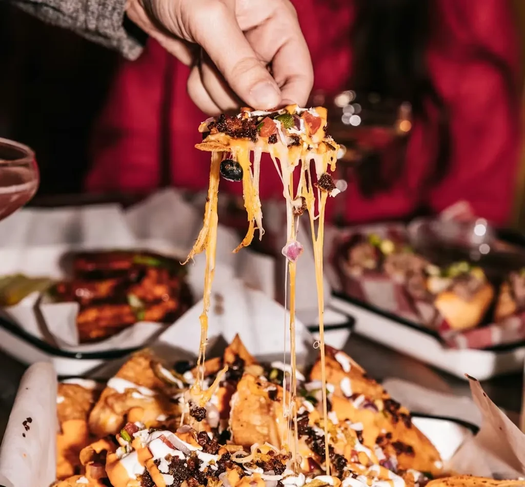 Hand pulls cheesy nacho from large dish with toppings.