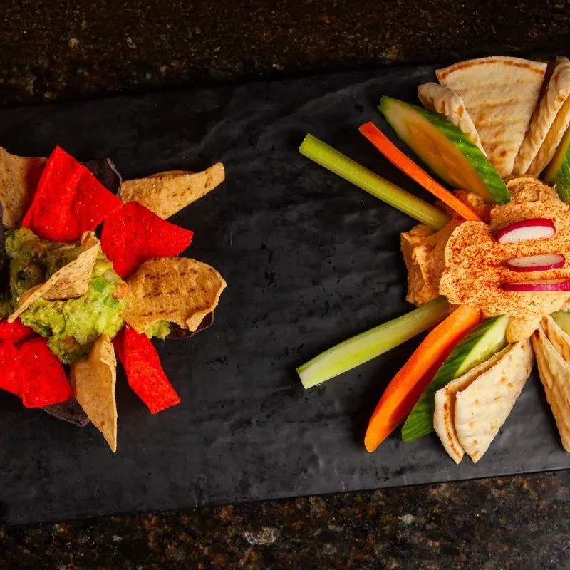 Guacamole and hummus platters with chips and vegetables on a board.