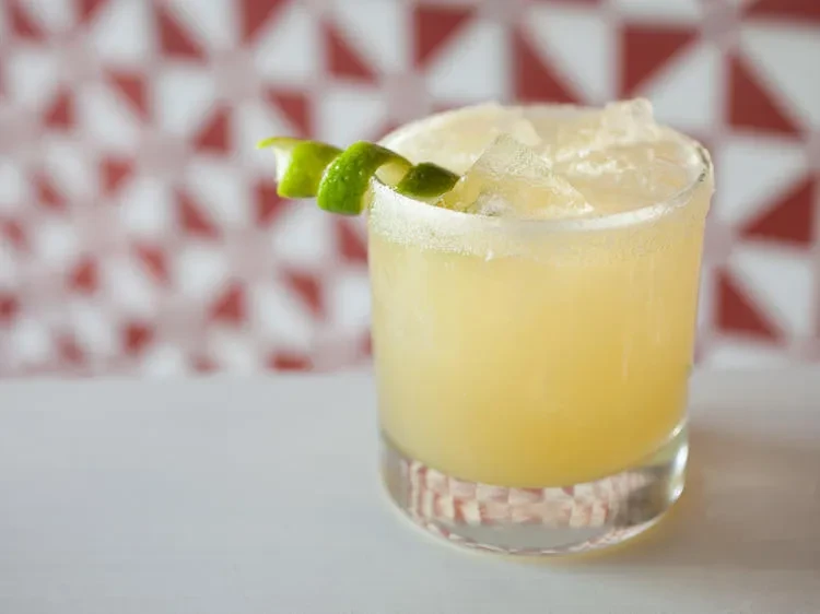 Pale yellow cocktail with crushed ice, salted rim, and lime twist.