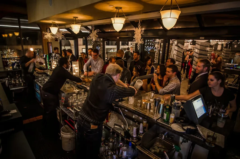 Nighttime bar scene with bartenders serving drinks and patrons socializing.