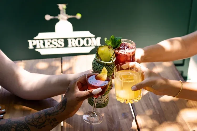 Four hands toast cocktails with "PRESS ROOM" sign in background.
