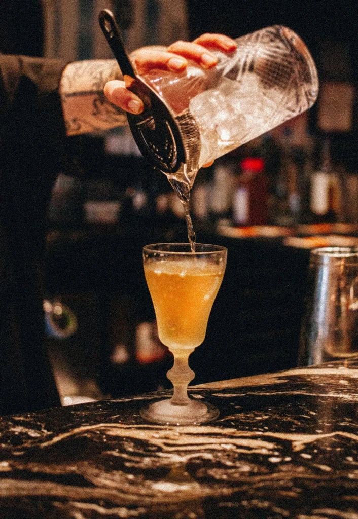 Bartender pours yellowish-orange liquid from a mixing glass into a frosted glass.