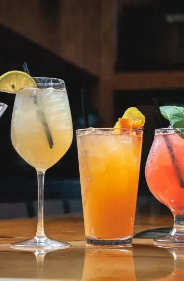 Three colorful drinks with straws and garnishes on a reflective surface.