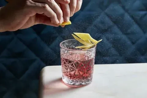 Hand squeezing lemon twist over a cocktail in a crystal glass.